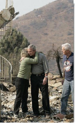 President George W. Bush hugs Kendra Jeffcoat as her husband Jay stands by in the midst of the charred rubble of their Rancho Bernardo, California home. The President met the couple during his visit Thursday, Oct. 25, 2007, to the Southern California areas ravaged by recent wildfires. White House photo by Eric Draper